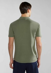 Uomo di spalle che indossa una polo a maniche corte verde oliva e pantaloni blu scuro su uno sfondo uniforme.