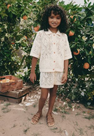 Copil mic cu păr creț stă desculț într-un costum tricotat crem lângă pomi de portocal și lăzi de lemn cu fructe în aer liber.