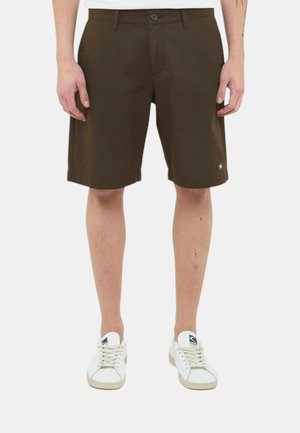Uomo che indossa pantaloncini marrone scuro fino al ginocchio e sneakers bianche, in piedi su uno sfondo chiaro uniforme.