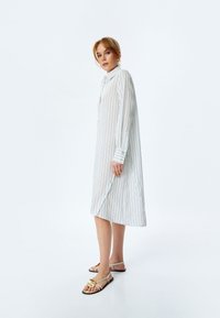 Vestito a camicia a righe bianche con maniche lunghe, polsini con bottoni e spacco laterale. La modella indossa sandali con dettagli dorati, in piedi su uno sfondo neutro.
