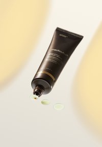 Brun tube med medicube Deep Vita Retinol Serum med gulddetaljer, hvor to små gulliggrønne dråber presses ud på en lys baggrund.