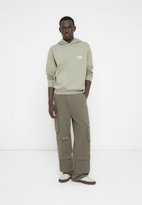 Φούτερ με κουκούλα σε ελαιί πράσινο χρώμα με τσέπη μπροστά και λογότυπο, συνδυασμένο με φαρδιά παντελόνια cargo με πολλές τσέπες και υφή.