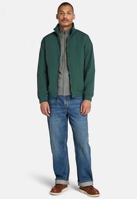 Grön zip-up-jacka med ståkrage, lager-på-lager över en grå zip-up-tröja. Bärs med ljusblå jeans och bruna skor.