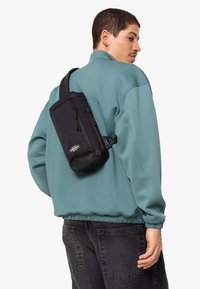 Borsa a tracolla nera con texture liscia, dotata di due scomparti con zip e un logo. Indossata sulla schiena sopra un felpa teal.
