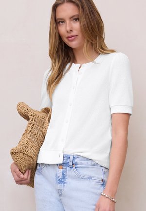 Femme portant un haut blanc à manches courtes boutonné et un jean bleu clair, tenant une pochette beige tissée dans sa main gauche.