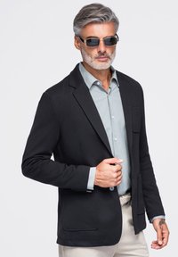 Homme mature aux cheveux gris et barbe portant un blazer noir, une chemise bleu clair, des lunettes de soleil et un pantalon de couleur claire sur un fond uni.