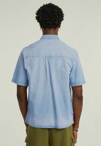 Ljusblå kortärmad skjorta i denimtyg, med standardkrage, en plym på baksidan och sydda detaljer nära fållen.