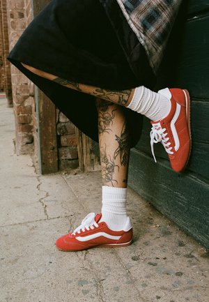 Nogi z kwiatowymi tatuażami, noszące białe skarpetki i czerwone trampki, jedna noga uniesiona do czarnej drewnianej ściany, na zewnątrz na betonowej powierzchni.