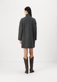 Kullsort oversized ullkåpe med høy krage, fulle ermer og rett snitt, paret med brune spisse cowboy boots.