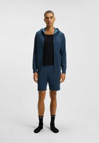 Felpa con zip di colore blu navy con cappuccio teal a contrasto, abbinata a pantaloni corti coordinati. Una canotta nera e calze nere completano l'outfit. Texture liscia.