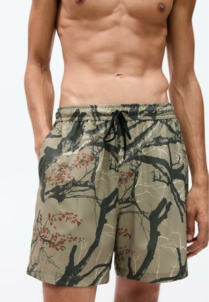 Shorts de bain camouflés en vert olive avec des motifs de feuilles noirs et rouges. Taille élastique avec cordon de serrage. Tissu léger et lisse.