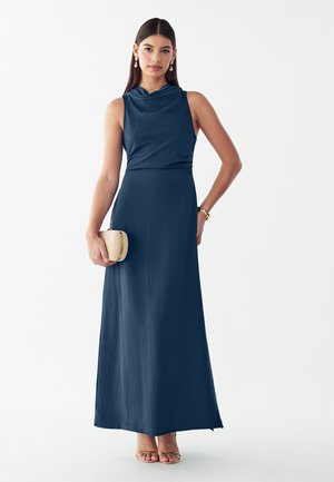 Frau trägt ein ärmelloses, bodenlanges dunkelblaues Kleid, hält eine kleine beige Clutch, mit langen dunklen Haaren und goldenen Schmuck.