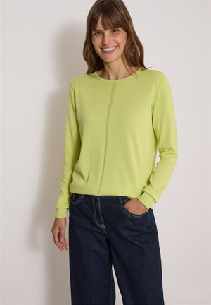 Smilende kvinde med brunt hår iført en lysegrøn langærmet sweater og mørkeblå jeans, stående med den ene hånd i lommen.