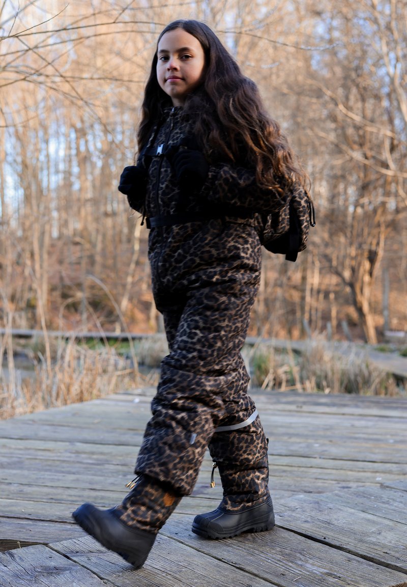 Leopard-print insulated snowsuit with a zipper front and pockets, paired with black waterproof boots. Background features wooden planks and foliage.