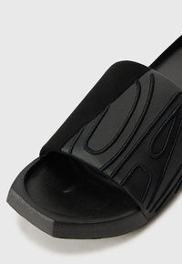 Sort slip-on sandal med struktureret overdel, der har hævede sorte stingmønstre, rektangulært tådesign og en mat finish på sålen.