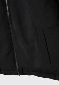 Section d'une veste noire montrant la fermeture éclair avant et une poche latérale rectangulaire avec des coutures visibles sur le tissu.