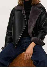 Veste en cuir noir avec col et doublure en fausse fourrure grise. Dotée d'une fermeture éclair et de manches larges. Le modèle porte un jean bleu foncé.