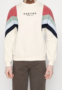Felpa crema con maniche a blocchi di colore rosso, verde, blu e blu navy. Presenta un collo rotondo e polsini a costine. Logo del marchio presente.
