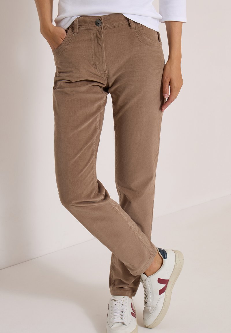 Personne portant un pantalon en velours côtelé marron et des baskets blanches avec des détails rouges et bleus, debout avec une jambe croisée et une main dans la poche.