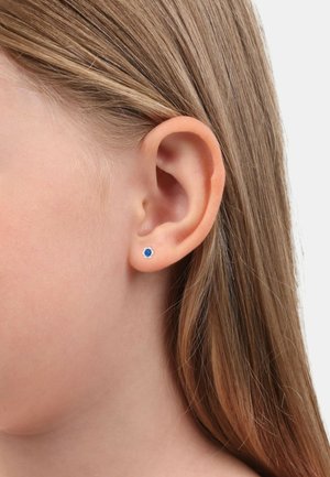 Pendientes de plata con una piedra preciosa azul en el centro, situados sobre un tono de piel claro y cabello largo y castaño.