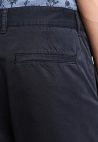 Navy cotton trousers with a back pocket and subtle stitching detail. Smooth texture with a fitted design. Floral pattern visible in background.