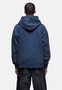 Felpa blu navy con cappuccio, realizzata in morbido tessuto, con una vestibilità rilassata e polsini a coste. Indossata con pantaloni in denim scuro.