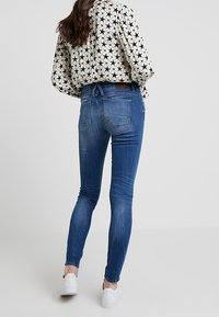 Sittande blå denimjeans med en lätt tvätt och bakfickor, kombinerade med en mönstrad svart och vit blus och vita sneakers.
