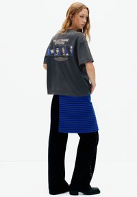 Tricou gri supradimensionat cu imprimeu grafic pe spate; asortat cu o fustă albastră cu dungi și pantaloni largi negri cu ținte argintii.