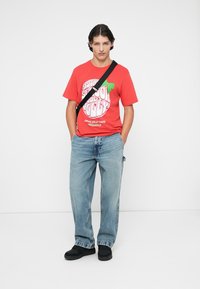 Crvena pamučna majica s bijelim grafičkim tekstom, plave traperice i crna obuća. Model pozira s crnom torbom preko ramena.