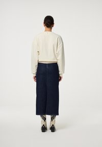 Persoon met donker haar in een bun, die een crèmekleurige sweatshirt, een donkerblauwe midi-denimrok en crèmekleurige enkellaarsjes met zwarte patronen draagt, met de rug naar de kijker gekeerd.