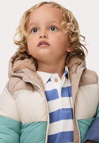 Vadderad jacka i beige, teal och brunt med en fluffig huva. Har en krage med knappar över en blå och vit randig skjorta.