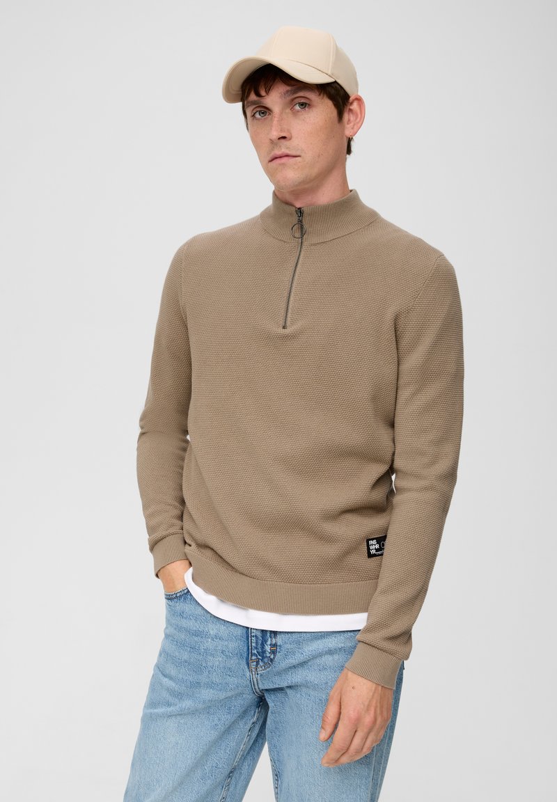 Beige Zip-up-Pullover aus strukturierter Strickware, mit langen Ärmeln und rundem Halsausschnitt. Kombiniert mit hellblauen Jeans und einer beigefarbenen Mütze.