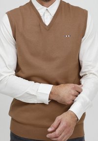 Brun stickad väst med V-hals och ribbad fåll, lagd över en vit skjorta. Har en liten flersidig logotyp på bröstet.