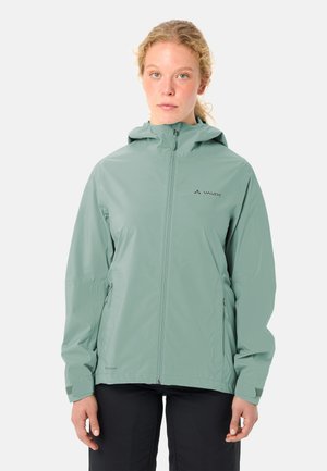 Femme portant une veste imperméable à capuche vert clair avec une fermeture éclair à l'avant et des poches latérales, debout devant un fond blanc uni.