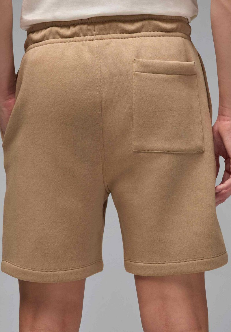 Shorts beiges décontractés avec taille élastique et une poche arrière, portés par une personne se tenant devant un fond uni.