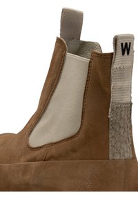 Botín de ante marrón con un panel lateral elástico beige, acento trasero texturizado gris y una pestaña de tirón con la letra "W" en negro.