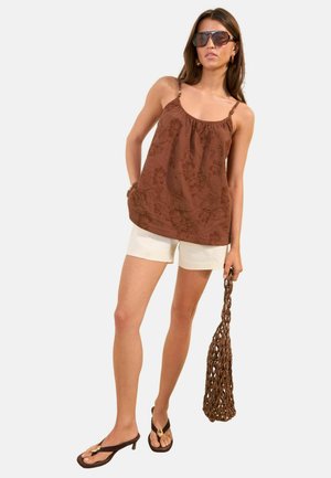 Naine, kes kannab pruunide lillemustriga varrukatuta topi, kreemikaid lühikesi pükse, tumedaid sandaale, päikeseprille ja kannab pruuni punutud kotti ühevärvilise tausta ees.