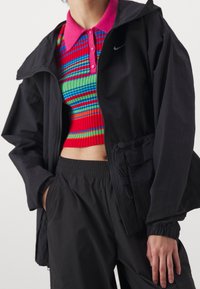 Chaqueta negra con cremallera y capucha sobre un crop top a rayas de colores que presenta patrones en rojo, verde y azul. Pantalones elásticos negros con cintura ajustable.