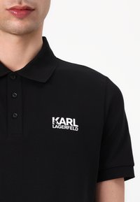 Zwarte polo met een omgeslagen kraag en drie knopen. Heeft een witte "KARL LAGERFELD" logo op het linker borstgebied. Glad katoen textuur.