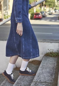 Osoba u traper haljini, bijelim čarapama i crnim tenisicama penje se uz vanjske betonske stepenice s rukom na ograde blizu ulice s parkiranim automobilima.