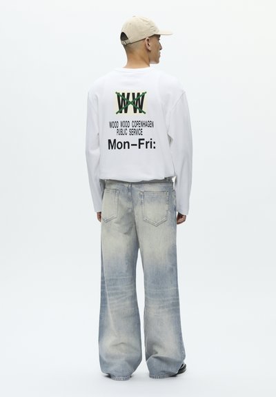 Muž v béžovej čiapke, bielom tričku s dlhými rukávmi a čiernym textom, a vyblednutých džínsach s širokými nohavicami stojaci pred jednoduchým bielym pozadím.