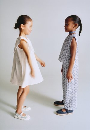Deux jeunes filles se font face ; l’une porte une robe blanche et des chaussures rayées, l’autre un haut à motifs et un pantalon, avec des chaussures foncées et un collier bleu.