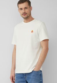 Weißes Baumwoll-T-Shirt mit einem kleinen bunten Flammenlogo auf der Brust. Rundhalsausschnitt und kurze Ärmel, kombiniert mit hellblauen Jeans.