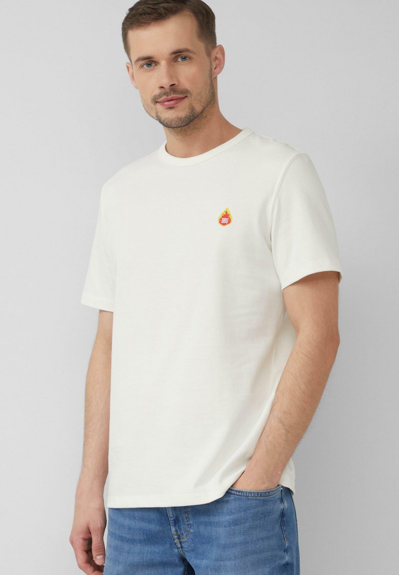 Weißes Baumwoll-T-Shirt mit einem kleinen bunten Flammenlogo auf der Brust. Rundhalsausschnitt und kurze Ärmel, kombiniert mit hellblauen Jeans.