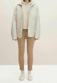 Chaqueta acolchada de color beige claro con dos bolsillos, llevada sobre un hoodie crema y unos jeans skinny marrón claro, combinada con zapatillas blancas.
