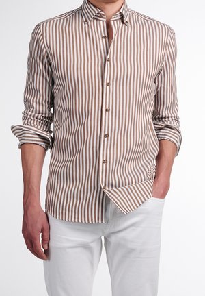 Hombre con camisa abotonada de rayas verticales beige y blancas, con las mangas remangadas y pantalones blancos, una mano en el bolsillo.