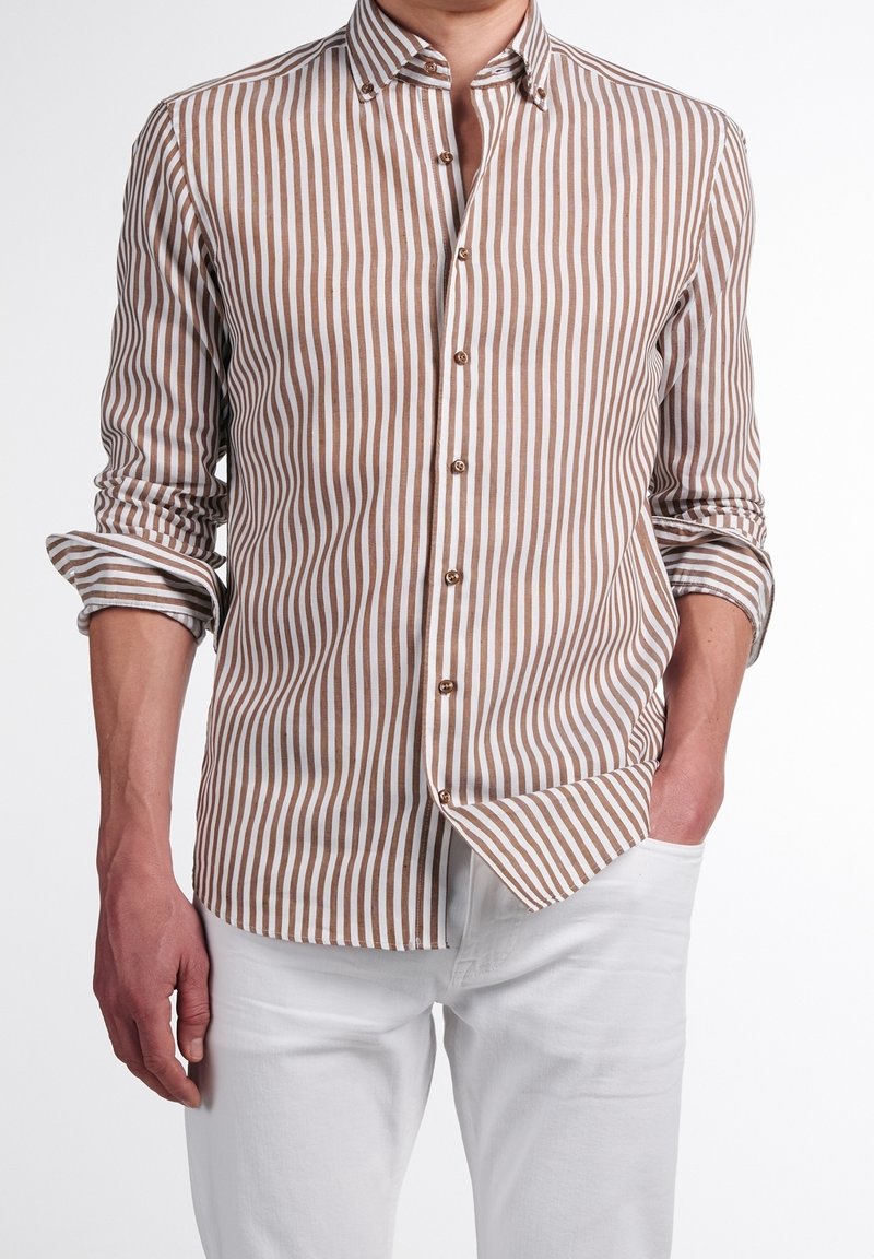 Homme portant une chemise beige et blanche à rayures verticales avec manches retroussées et pantalon blanc, une main dans la poche.