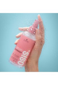 Un flacon pulvérisateur rose avec un couvercle texturé, tenu dans une main. Des gouttes d'eau sur la bouteille créent une apparence humide et rafraîchissante sur un fond bleu.