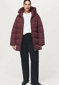 Chaqueta de plumas burdeos con capucha, con un diseño acolchado y cierre de cremallera. Combinada con una camisa blanca y jeans negros.