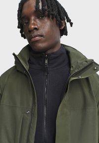 Olivově zelená vnější bunda s vysokým límcem, překrytá námořnickou fleecovou mikinou se zipem. Obsahuje zipy, patentky a hladkou texturu látky.